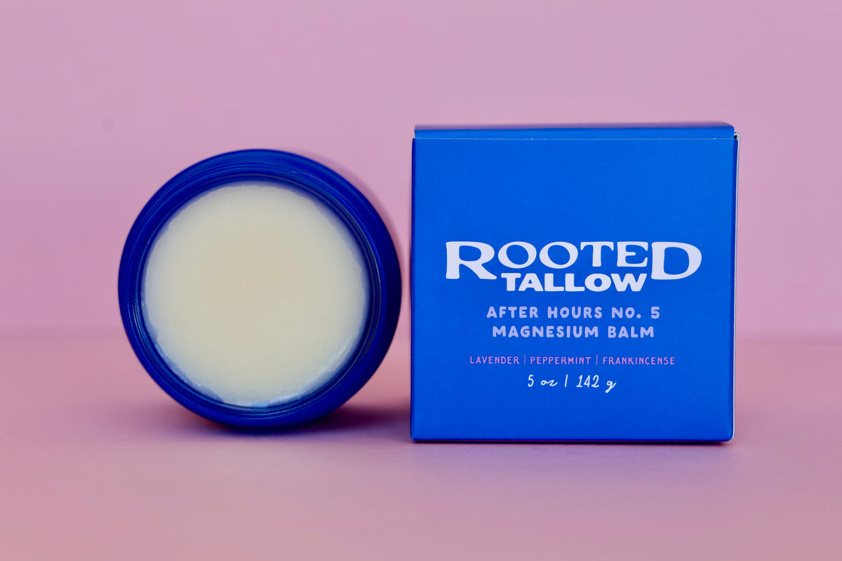 An open round jar of Rooted Tallow After Hours No. 5 Magnesium Balm next to its blue box on a solid pink background.